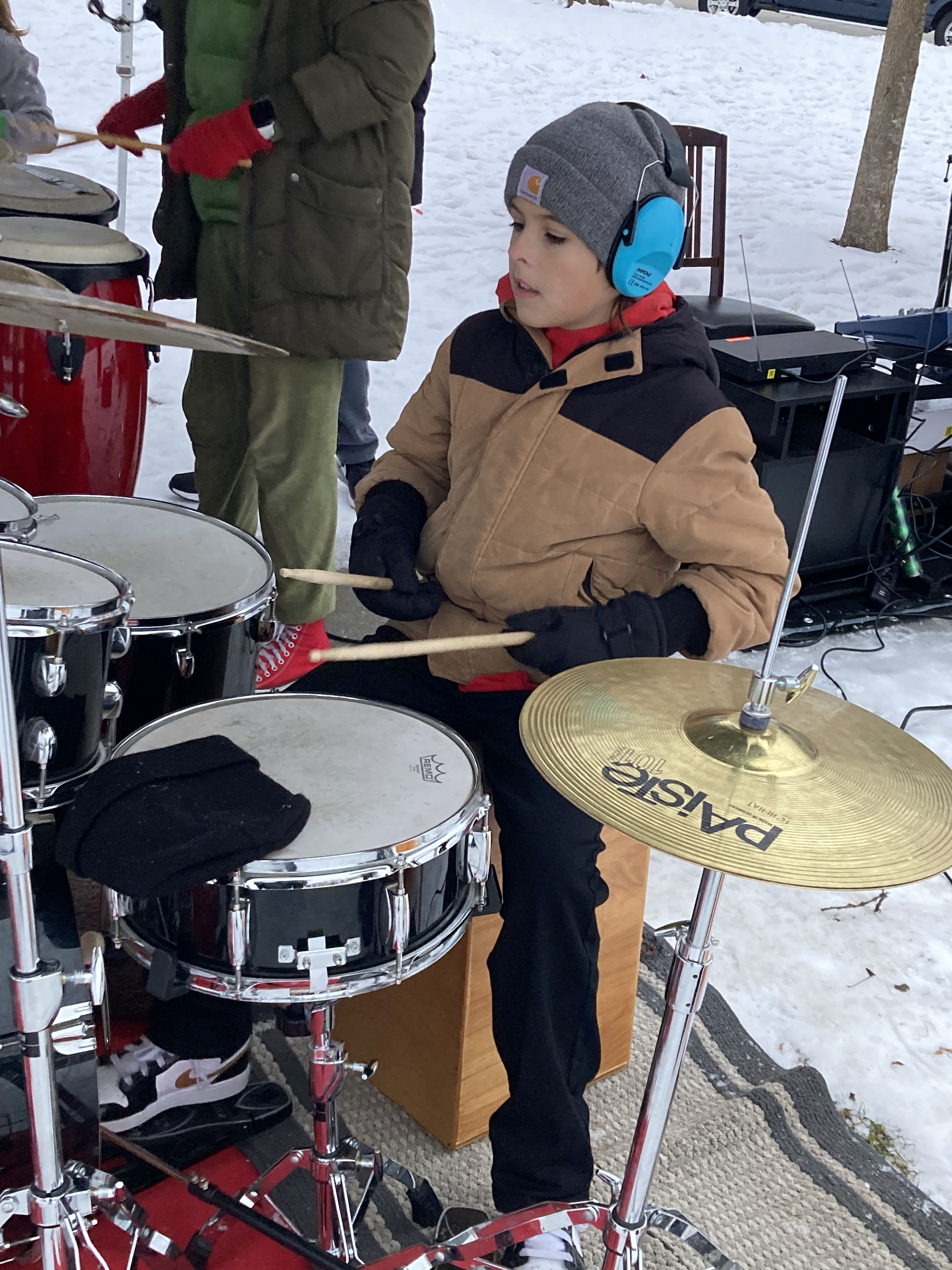 student in a tan jacket and grey hat with hearing protection at holiday event playing the drum kit