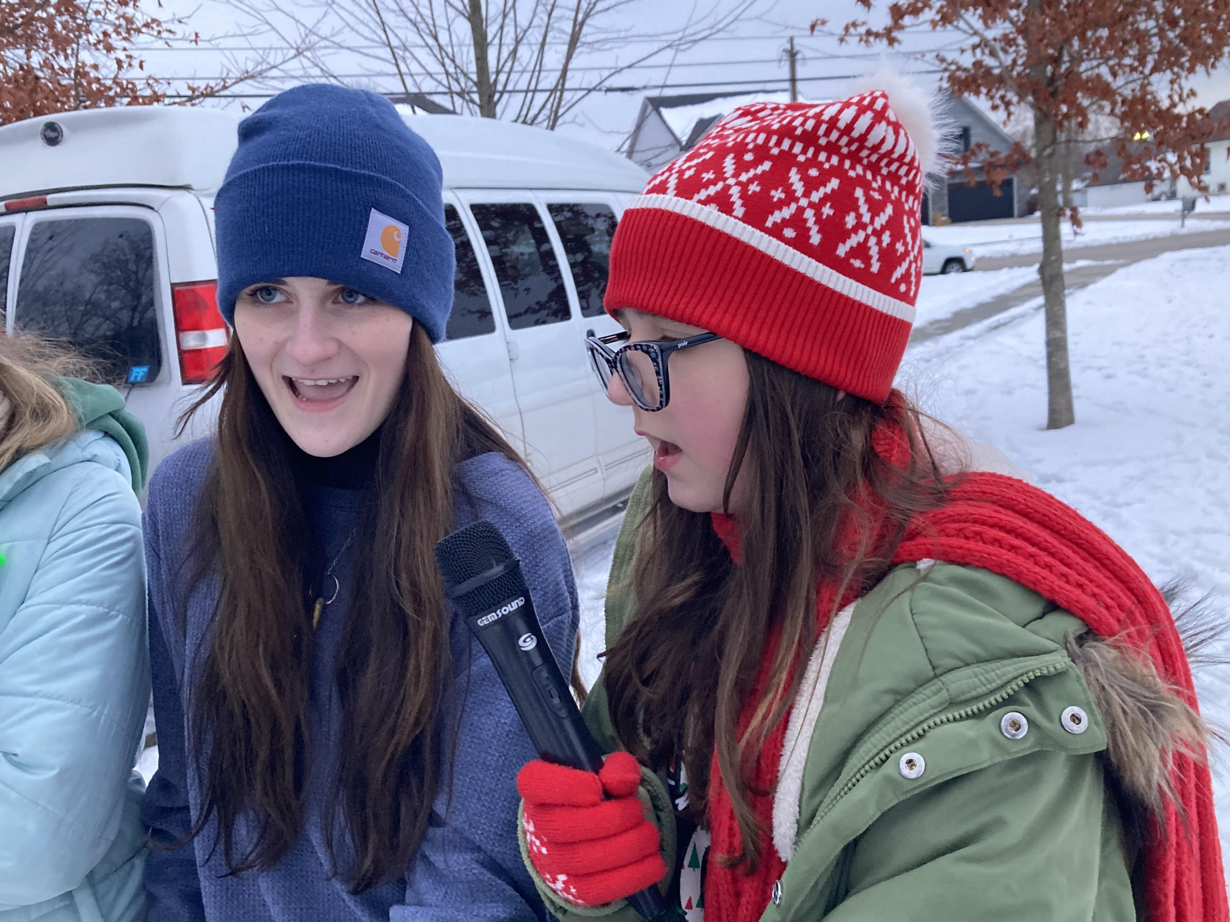 2 students singing at a holiday event with a microphone: one in a blue jacket and hat, one in a green jacket and red hat