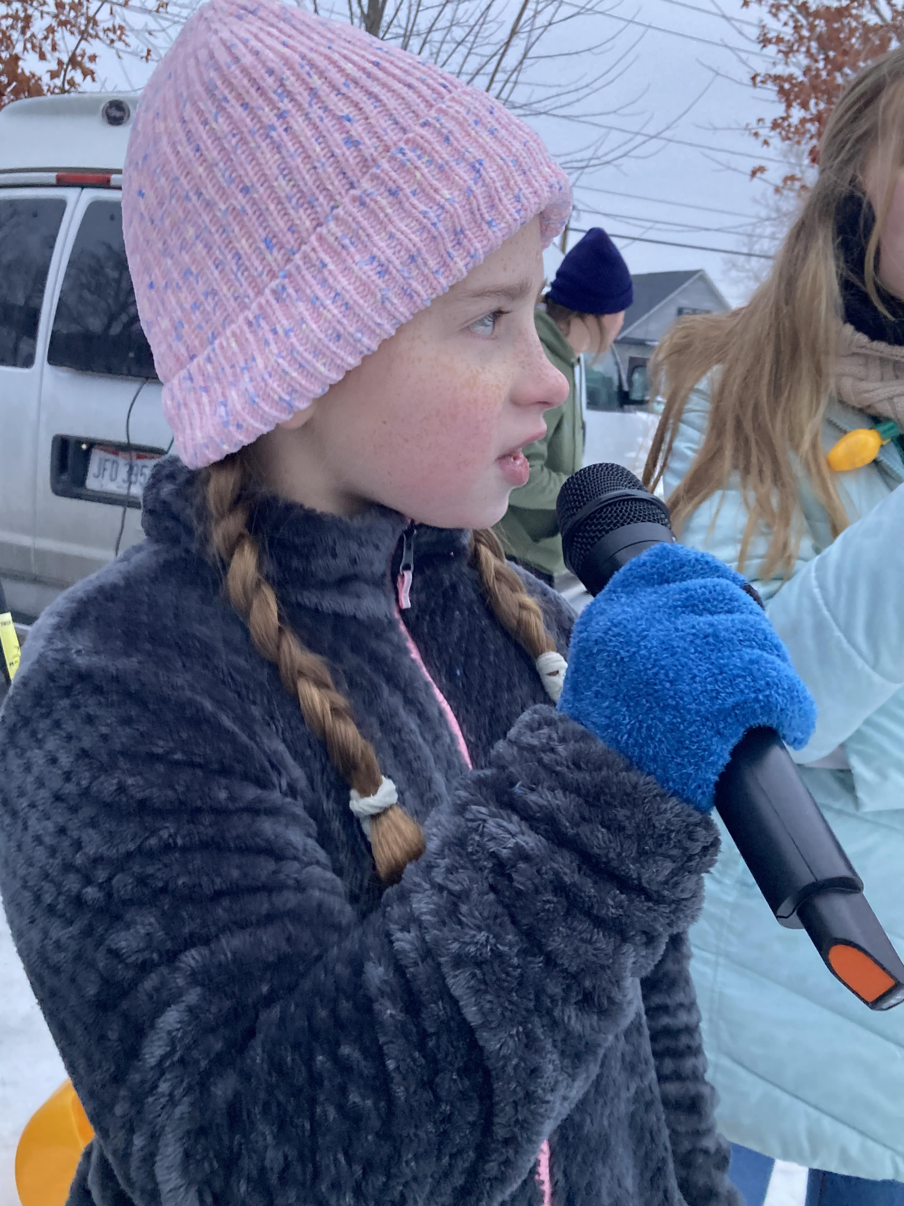student in a grey jacket and pink hat singing into a microphone at a holiday event