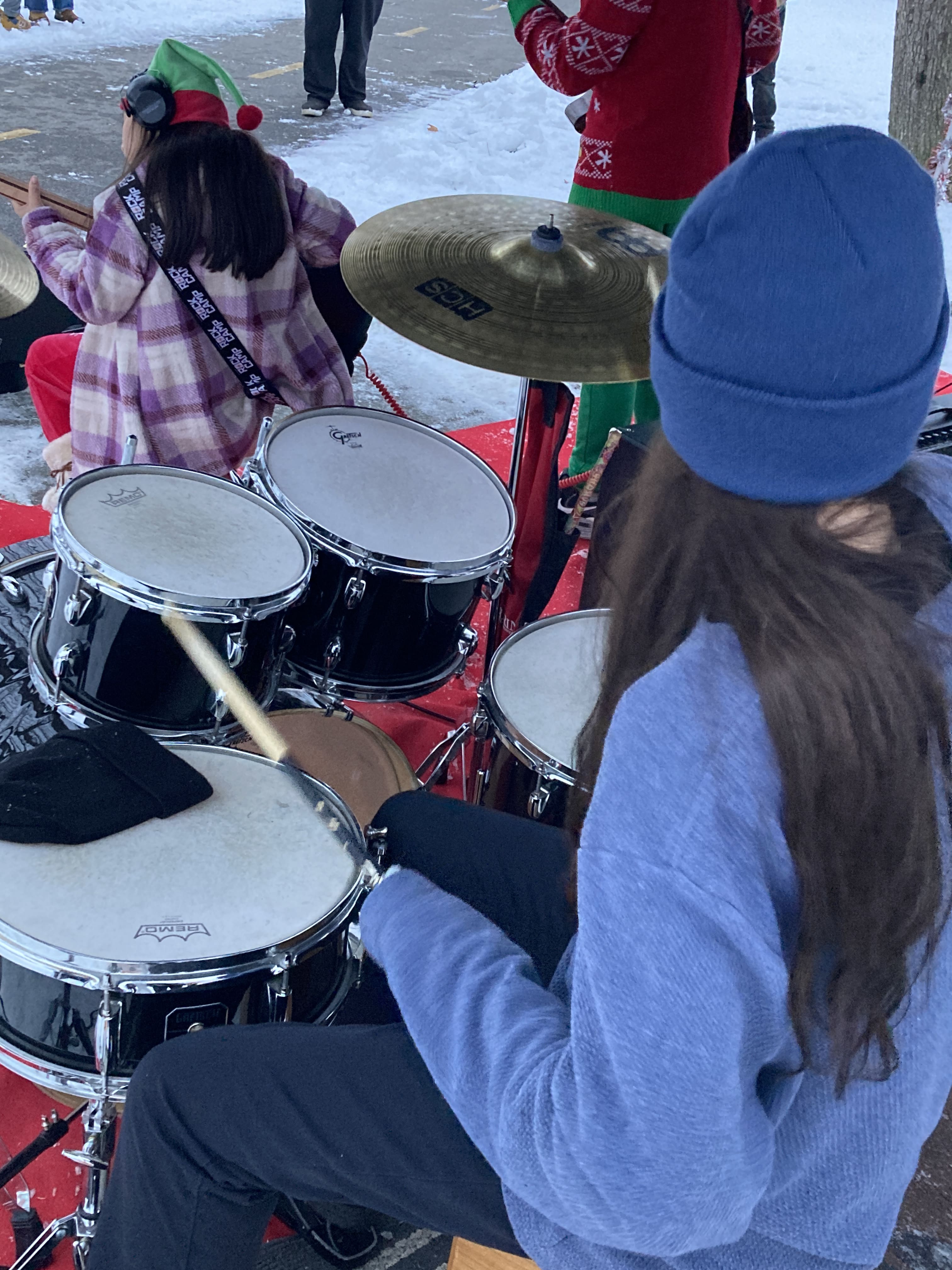 student in a blue jacket and hat playing the drum kit with hearing protection at a holiday event